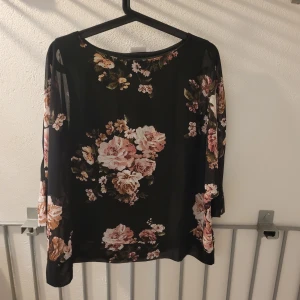 Svart blommig blus med vida ärmar - Säljer en svart blus från VILA med stora blommiga mönster i rosa, beige och grönt. Blusen har vida, halvtransparenta ärmar och en lös passform. Perfekt för dig som gillar romantiska vibes och vill ha något luftigt och snyggt.