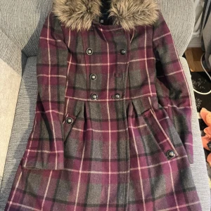 Rutig kappa med fuskpäls och bälte - Säljer en rutig kappa i mörklila och grå toner med snygg fuskpäls vid kragen. Kappan har dubbelknäppning, bälte i midjan och dekorativa knappar. Perfekt för dig som vill ha en statement-jacka med vintagevibbar.