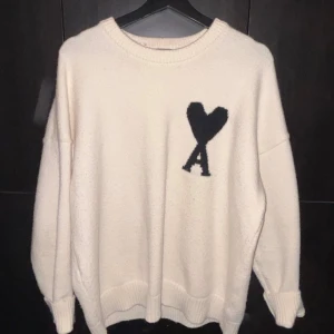 Vit stickad tröja från Ami Paris - Säljer en vit stickad tröja från Ami Paris med svart hjärta och bokstaven A broderat på bröstet. Tröjan har rund hals, ribbade muddar och en loose passform. Perfekt för dig som gillar stilrena och trendiga plagg. Storleken är M, finns en enda liten defekt längst ner på tröjan som knappt är märkvärdig