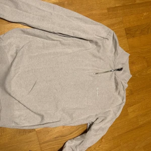 Ljusgrå/Beige half zip tröja Bondelid - Ljusgrå långärmad tröja från Bondelid med broderad logga på bröstet och half zip-dragkedja vid halsen. Tröjan har ribbade muddar och är tillverkad i mjuk bomull, perfekt för chill dagar.