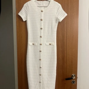 Vit kortärmad klänning med knappar - Stilren vit klänning i strukturerat tyg med korta ärmar och dekorativa guldknappar framtill. Modellen är knälång med slits nertill och har två fickdetaljer vid höften. Dragkedja i ryggen för enkel påklädning. Perfekt för dig som gillar klassisk och chic stil.