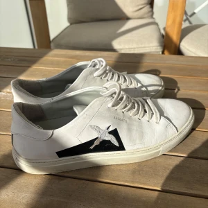 Vita sneakers från Axel Arigato - Snygga vita sneakers från Axel Arigato i skinn med klassisk låg profil och rund tå. Skorna har snörning och en broderad fågel på sidan i svart och vitt, samt diskret Arigato-logga. Perfekta för en clean och stilren look.