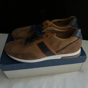 Bruna skor från Paul Hunter - Snygga skor från Paul Hunter i olika bruna nyanser med mörkblå detaljer och snörning. Skorna har en mix av mocka och skinn, vit sula och mörkblå ränder på sidan. Perfekta för dig som gillar en klassisk look