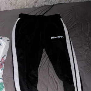 Palm Angels svarta mjukisbyxor med vita ränder - Svarta mjukisbyxor från Palm Angels med vita ränder längs sidorna och logga tryckt på låret. Byxorna har dragsko i midjan och är tillverkade i ett glansigt syntetmaterial. Perfekt för en streetwear-look.