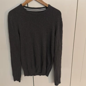 Mörkgrå stickad tröja med rund hals - En stilren mörkgrå stickad tröja med rund halsringning och långa ärmar. Perfekt för lager-på-lager och funkar till både jeans och chinos. Tröjan har en klassisk passform och ribbade muddar vid ärmslut och nederkant.
