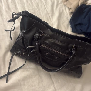 Banlenciaga liknande väska!! - Cool svart handväska i skinn med silverfärgade detaljer och dragkedja. Väskan har dubbla handtag, dekorativa nitar och en praktisk ytterficka. Perfekt för skolan!! Har lite defekter, har tagit bilder på de! Men jag tycker inte att man tänker på de!🥰🥰