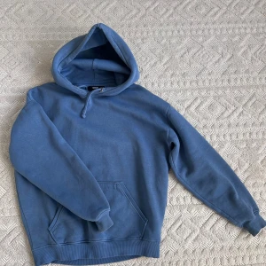 Blå hoodie med huva och ficka - Säljer en blå hoodie från Bik Bok med stor huva och känguruficka framtill. Tröjan har ribbade muddar vid ärmslut och nederkant samt snörning i huvan. Perfekt för chill dagar eller när du vill vara extra bekväm.