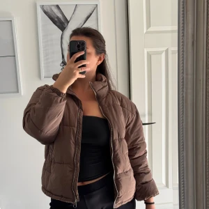 Brun pufferjacka från Pull&Bear - Säljer en brun pufferjacka från Pull&Bear med hög krage och dragkedja framtill. Jackan är quiltad och har en loose passform, perfekt för kyligare dagar. Fodrad insida och elastiska muddar vid ärmslut. Enkel och stilren design.