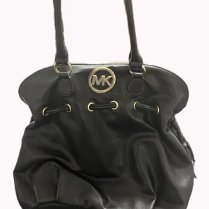 Svart handväska Michael Kors skinn - Svart handväska från Michael Kors i mjukt skinn med snygga guldfärgade detaljer och MK-logga framtill. Väskan har snörningsdetalj upptill och långa handtag, perfekt för dig som gillar klassisk och stilren design. 📦 Skickar snabbt 🐶 Hem med djur (kan förekomma lite päls) 💬 Fråga gärna om fler bilder eller paketpris  