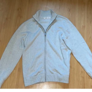 Dobber tröja  - Säljer en stilren ljusgrå stickad kofta från Dobber i storlek M. Koftan har hel dragkedja framtill, ribbade muddar och hög krage. Perfekt att slänga på sig över en t-shirt för en clean look.