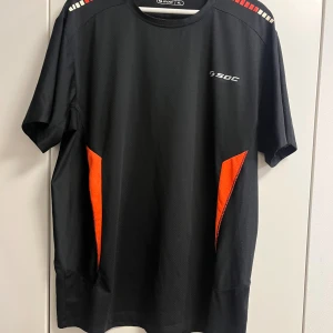 Svart tränings-tshirt från SOC XL - Svart tränings-tshirt från SOC i funktionsmaterial med orange detaljer på sidorna och axlarna. Kortärmad modell med rund hals och diskret logga på bröstet. Perfekt för dig som vill ha en sportig och bekväm look.