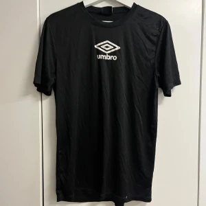 Svart Umbro t-shirt med logga - Svart t-shirt från Umbro med vit logga tryckt på bröstet. Klassisk passform och rund halsringning. Tillverkad i mjukt material som känns skönt mot huden. Perfekt för dig som gillar sportiga och stilrena plagg.