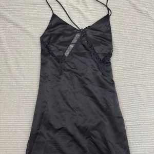 Svart långklänning i satin från H&M - Supersnygg silvrig långklänning i satin från H&M med tunna axelband och spetsdetaljer vid bysten. Klänningen har öppen rygg med snörning. 