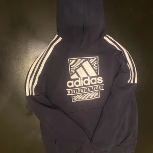 Mörkblå Adidas hoodie med tryck - Mörkblå hoodie från Adidas med klassiska vita ränder längs ärmarna och stort Adidas Worldwide Sport-tryck på bröstet. Tröjan har huva och är tillverkad i mjuk bomullsmix. Perfekt för en sportig och avslappnad stil.