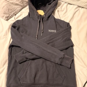 Morris hoodie - Mörk blå hoodie från Morris Sporting Club med broderad logga på bröstet. Klassisk modell med huva och snörning, känguruficka framtill och ribbade muddar. Tillverkad i mjuk bomull, perfekt för chill dagar eller när du vill ha en clean look.