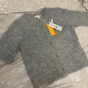 Grå kofta med pärlknappar från Gina Tricot - Superfin ljusgrå kofta från Gina Tricot med kort ärm och snygga pärlknappar framtill. Koftan är stickad och har en rund halsringning med ribbade kanter. Perfekt att slänga över en topp. Strl 170 motsvarande xs/s