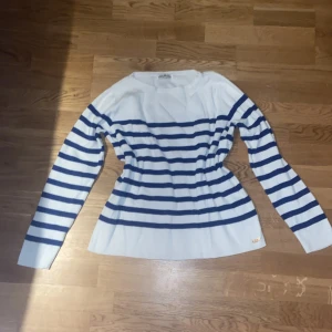 Randig tröja från Hampton Republic - Vit och marinblå randig tröja från Hampton Republic med lång ärm och klassisk båtringad hals. Tröjan är stickad i bomull och har en liten metallbricka nertill som snygg detalj. Perfekt för en stilren och avslappnad look.