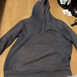 Mörkgrå hoodie från Hoodrich - Säljer en mörkgrå hoodie från Hoodrich med stor logga på ryggen och mindre tryck på bröstet. Tröjan har huva med snörning och en stor magficka. Perfekt för dig som gillar streetwear och vill ha en chill look.