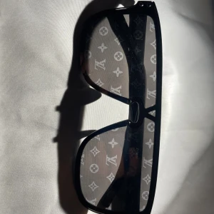 Louis Vuitton svarta solglasögon - Snygga svarta solglasögon från Louis Vuitton med bred båge och rektangulär form. Glasen har det ikoniska LV-monogrammet tryckt över hela ytan för en riktigt lyxig vibe. Perfekta för dig som vill sticka ut och visa stil.