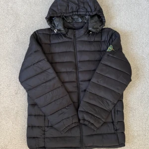 Svart pufferjacka från Stone Island - Svart pufferjacka från Stone Island med avtagbar huva och klassisk patch på ärmen. Jackan har horisontella sömmar, dragkedja framtill och elastiska muddar vid ärmslut. Perfekt för kalla dagar och riktigt snygg streetstil. Storlek L men sitter som M.