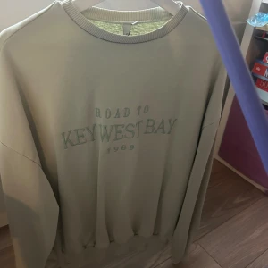 Ljusgrön oversized tröja  - Ljusgrön oversized tröja från NELLY med broderad text 'Road to Key West Bay 1989' framtill. Tröjan har rund hals, ribbade muddar och lång ärm. Perfekt för en chill och avslappnad stil.