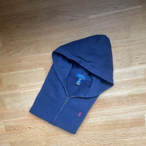 Marinblå hoodie från Polo Ralph Lauren - Snygg marinblå hoodie från Polo Ralph Lauren med dragkedja och huva. Tröjan har det klassiska röda Polo-logot broderat på bröstet, två fickor fram och ribbade muddar. Perfekt för chill dagar och enkel att matcha med jeans eller joggers.   Storlek XL kids=Storlek S men 