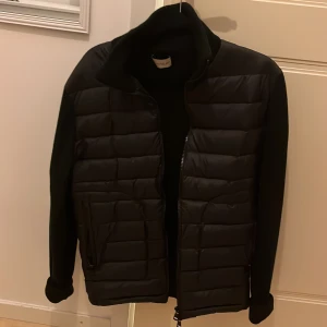 Svart quiltad jacka från Moncler - Svart Moncler jacka med quiltad front och ärmar i stickad kvalitet. Jackan har hög krage, dragkedja framtill och Moncler-logga på ena ärmen. Snygg och stilren design som passar perfekt till höst och vinter. Använd 2-3 gånger, är i nyskick!  