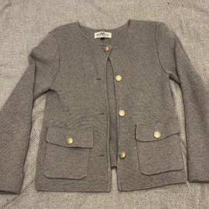 Grå kofta från Hampton Republic -  Grå kofta från Hampton Republic med snygga guldknappar och två stora fickor framtill. Jackan är stickad i mjukt material och har rund halsringning samt lång ärm. Köpt för 700kr