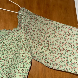 Grön blommig kortklänning från H&M storlek XL - Supersöt ljusgrön topp från H&M med små röda och rosa blommor. Toppen har  långärmade, luftiga ärmar och smockad överdel med volangdetaljer vid halsen och nederkant. Tillverkad i mjukt polyester-tyg, perfekt för sommarens alla tillfällen.