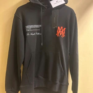 AMIRI hoodie, helt ny.  - Svart hoodie från AMIRI med stor ficka framtill, huva med snörning och coolt tryck i vitt och rött både fram och bak. Materialet är 100% bomull och tröjan har en relaxed passform med långa ärmar. Snygg LA-vibe och streetstyle känsla. Skriv önskad storlek efter lagd order. 