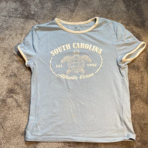 Ljusblå South Carolina t-shirt H&M - Ljusblå t-shirt från H&M med vit text och sköldpaddsgrafik på bröstet. Texten säger 'South Carolina Atlantic Ocean Est. 1992'. T-shirten har vita kontrastkanter vid ärmslut och halsringning. Tillverkad i mjuk bomull och har en klassisk passform med korta ärmar.