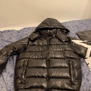 Svart Moncler pufferjacka med huva - Säljer en svart pufferjacka från Moncler med avtagbar huva och dragkedja framtill. Jackan har vita detaljer på ena ärmen och klassisk Moncler-logga. Fodrad och riktigt varm, perfekt för kalla dagar. Snygg, glansig finish och två fickor med dragkedja. Har använt en gång i skolan tills jag märkte att den var för stor 