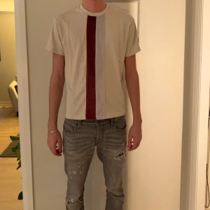 Vit t-shirt med röd sammetsrand La Vichy - Vit t-shirt från La Vichy i storlek M med en bred röd sammetsrand och en smalare vit rand framtill. Klassisk rund halsringning och korta ärmar. Snygg detalj med svart patch på ena ärmen. Perfekt för dig som vill sticka ut lite extra.