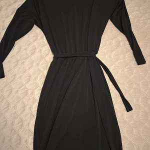 Svart långärmad omlottklänning - Svart långklänning med långa ärmar och omlottknytning i midjan. Klänningen har en enkel och stilren design med ett avtagbart bälte som markerar midjan. Perfekt för dig som gillar minimalistisk och tidlös stil.