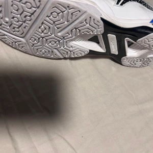 Li-Ning vita träningsskor - Snygga vita träningsskor från Li-Ning med svarta och blå detaljer samt röda och blå öglor för snörningen. Skorna har ventilerande meshpartier, coola grafiska mönster på sidan och slitstark sula med grymt grepp. Perfekta för dig som gillar sport och vill sticka ut lite extra. Med en exstra tabil sula badminton skor men funkar för alla anan sport i hallar 