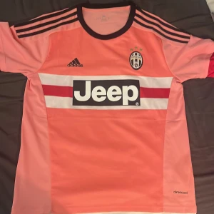 Juventus rosa fotbollströja Adidas - Juventus matchtröja från Adidas i rosa med svarta detaljer och vita/röda ränder över bröstet. Jeep-logga på framsidan, Juventus klubbmärke med tre stjärnor och Adidas-logga. Kortärmad, rund hals och tillverkad i lätt, ventilerande polyester (climacool). Pris kan diskuteras