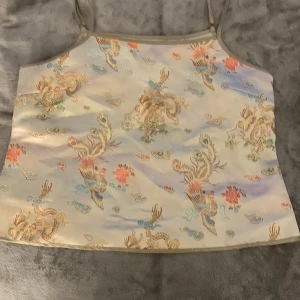 Beige sidenlinnetopp från NA-KD - Säljer en beige linnetopp från NA-KD i siden med asiatiskt inspirerat broderat mönster i guld, blått, rött och grönt. Toppen har smala axelband och en croppad passform. Ge gärna prisförslag! Jag har köpt den från Sellpy för ett tag sedan och har själv aldrig användt!