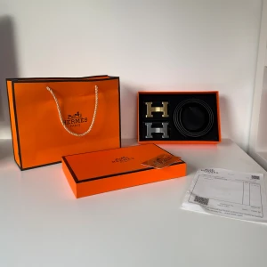 Svart H bälte med H-spänne - Exklusivt svart bälte från H i slätt skinn med två utbytbara H-spännen i guld och silver. Bältet levereras i original orange box med certifikat och kvitto. Perfekt accessoar för dig som vill ha en lyxig och stilren look.