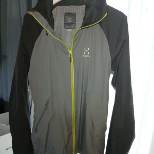 Grå och svart vindjacka från Haglöfs - Snygg vindjacka från Haglöfs i grått och svart med huva och gul dragkedja. Jackan har lång ärm, två fickor framtill och är tillverkad i lätt syntetmaterial. Perfekt för utomhusaktiviteter och har en sportig look.