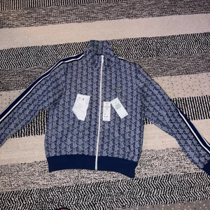 Blå mönstrad track jacket från Lacoste - Snygg blå track jacket från Lacoste med vitt mönster, vita ränder längs ärmarna och dragkedja framtill. Jackan har hög krage, ribbade muddar och klassisk Lacoste-logga på bröstet. Kvitto finns, köpt i Lacoste affär.