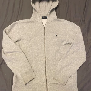 Grå hoodie från Polo Ralph Lauren - Snygg ljusgrå hoodie från Polo Ralph Lauren med dragkedja och klassisk broderad logga på bröstet. Tröjan har huva, långa ärmar och två fickor framtill. Perfekt för chill dagar eller när du vill ha en enkel och stilren look. Liten defekt vid vänster arm (se sista bild) 