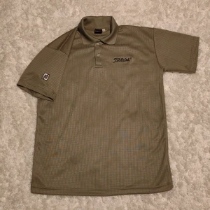 Beige Titleist piké i storlek L - Snygg beige pikétröja från Titleist med diskret rutmönster och broderad logga på bröstet. Tröjan har korta ärmar, klassisk krage och knappar framtill. Perfekt för golfbanan eller chill dagar. Materialet är lätt och andas, troligen polyester.