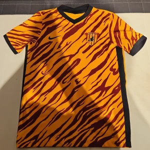 Benevento Calcio tigertröja Nike XL - Säljer en Benevento Calcio fotbollströja från Nike i XL med tigerinspirerat mönster i gult och vinrött. Tröjan har korta ärmar, svart V-ringad krage och är gjord i lätt Dri-Fit material som andas. Klubbmärket sitter på bröstet och Nike-loggan är tryckt på framsidan.