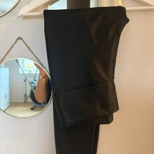 Svarta skinnimitation byxor - Säljer ett par svarta byxor i skinnimitation med stilren look. Byxorna har raka ben och en snygg söm vid knäna som ger extra edge. Perfekta för dig som vill ha en trendig och clean stil.