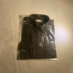 Svart skjorta med knappar - Stilren svart skjorta från Clique med klassisk krage och snygga svarta knappar längs framsidan och vid manschetterna. Skjortan har en clean look och är perfekt för dig som gillar enkel och tidlös stil. Materialet känns mjukt och bekvämt mot huden.