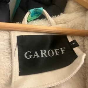 Beige teddyjacka med fuskpäls från Garoff - Säljer en beige teddyjacka från Garoff med stor huva och fluffiga fuskpälsdetaljer längs krage, ärmslut och öppning. Jackan har dragkedjefickor och en loose passform som ger en chill vibe. Perfekt för kalla dagar när du vill vara både varm och snygg.