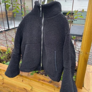 Svart teddyjacka från H&M, stl M - Mysig svart teddyjacka från H&M i storlek M. Jackan har hög krage, dragkedja framtill och två stora fickor. Perfekt för kyliga dagar och ger en cool, avslappnad vibe. Materialet är fluffigt och mjukt, vilket gör den extra skön att bära.