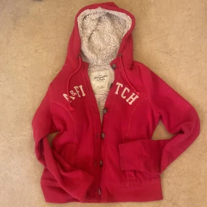 Röd hoodiejacka från Abercrombie & Fitch - Röd/rosa hoodiejacka från Abercrombie & Fitch med mysigt teddyfoder innuti Jackan har knappar framtill, dragsko i luvan och broderad logga på bröstet. Perfekt för kyliga dagar och riktigt skön att ha på sig.