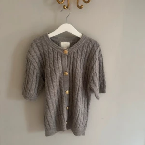 Grå kabelstickad kofta med knappar - Grå kabelstickad kofta med korta ärmar och snygga guldfärgade knappar framtill. Rund halsringning och ribbade muddar ger en chill och trendig vibe. Perfekt att slänga över en t-shirt för extra stilpoäng.