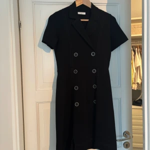 Svart kortklänning från Sandro - Snygg svart kortklänning från Sandro med dubbelknäppt design och korta ärmar. Klänningen har krage och sex dekorativa knappar framtill som ger en cool, dressad look. Perfekt för dig som gillar stilrena och moderna plagg. Nypris 2000 kr.